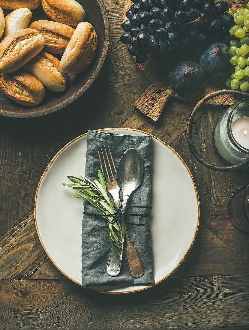 Tischgedeck mit Serviette, Besteck und Olivenzweigen daneben Windlicht, Semmeln und Obst