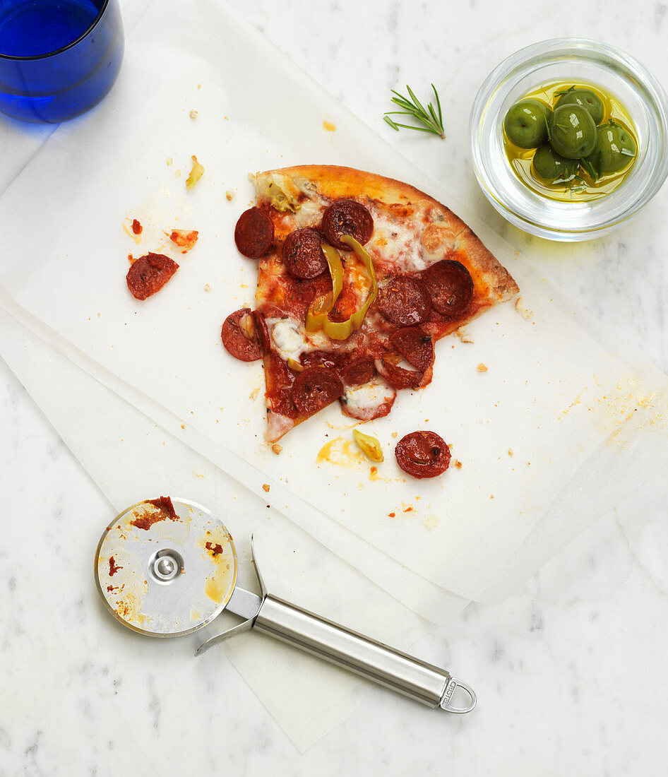Pizzastück mit Peperoniwurst, daneben grüne Oliven