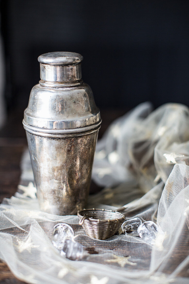 A silver cocktail shaker and bar sieve on a starry cloth