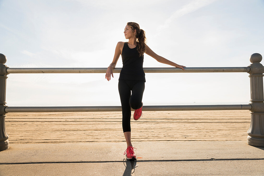 A young woman wearing sports clothes leaning against a railing on a beach