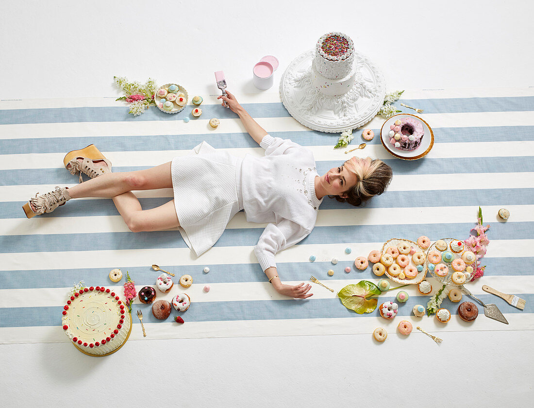 Frau mit Pinsel in der Hand, umgeben von Donuts und Kuchen, liegt auf dem Boden