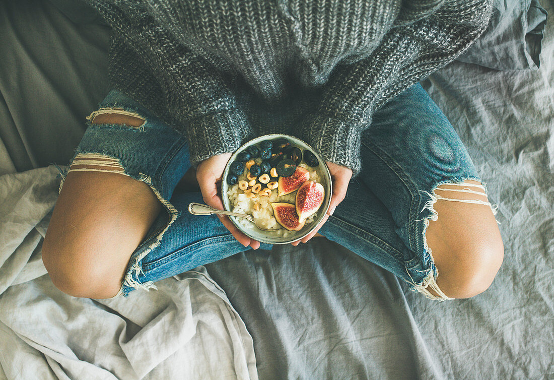 Healthy winter breakfast in bed: Woman in sweater and jeans holding rice coconut porridge with figs, berries, hazelnuts