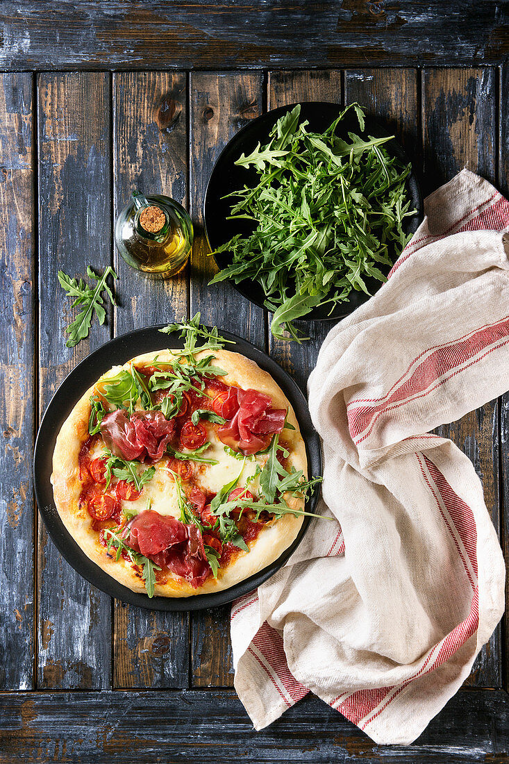 Whole homemade pizza with cheese and bresaola, served on black plate with fresh arugula, olive oil and kitchen towel