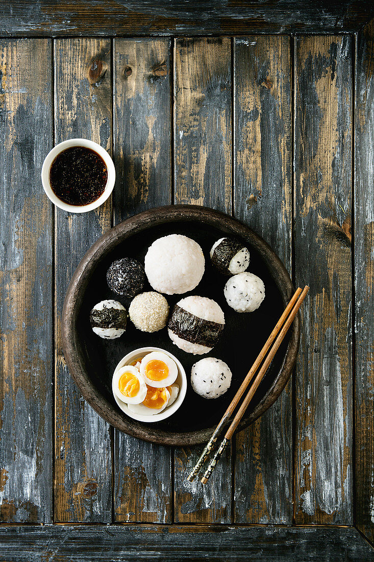 Stone tray with different size rice balls with black sesame and seaweed nori, served with soft boiled eggs, soy sauce