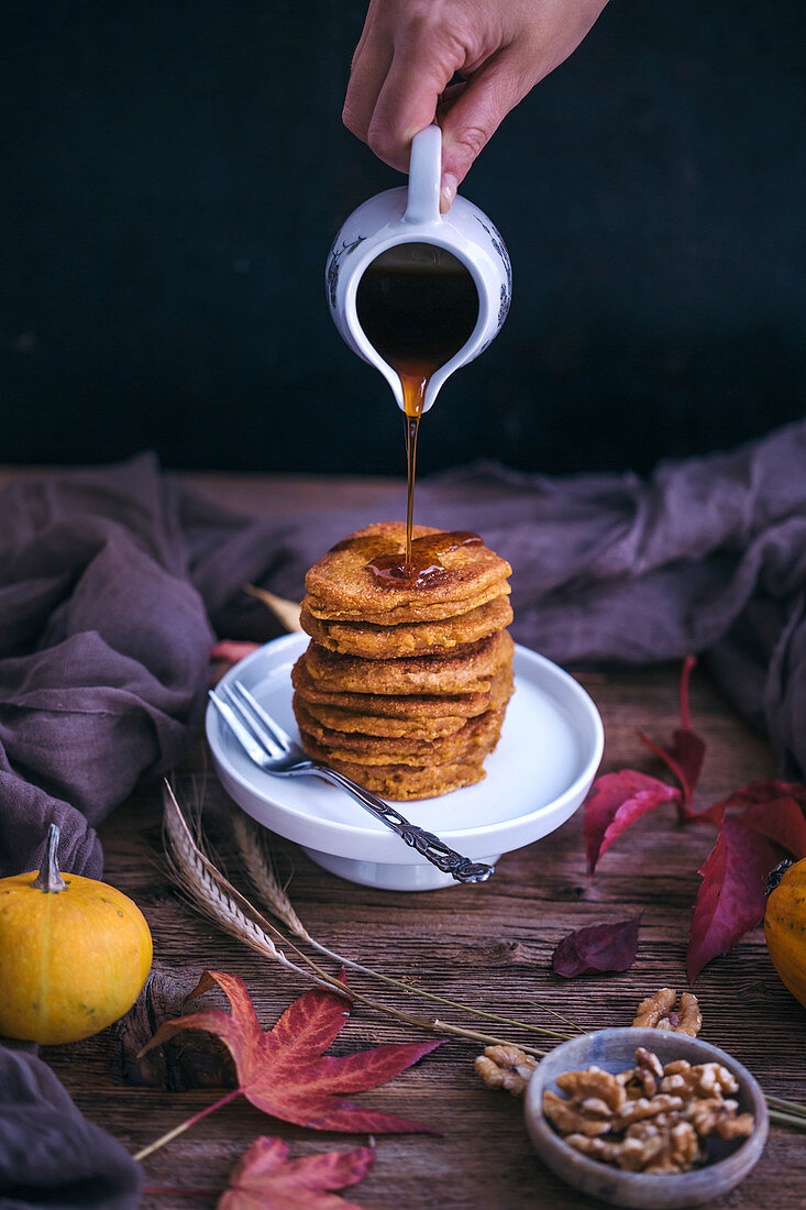 Woman pouring maple syrup on pumpkin pancakes served on a little cake stand on rustic wooden table