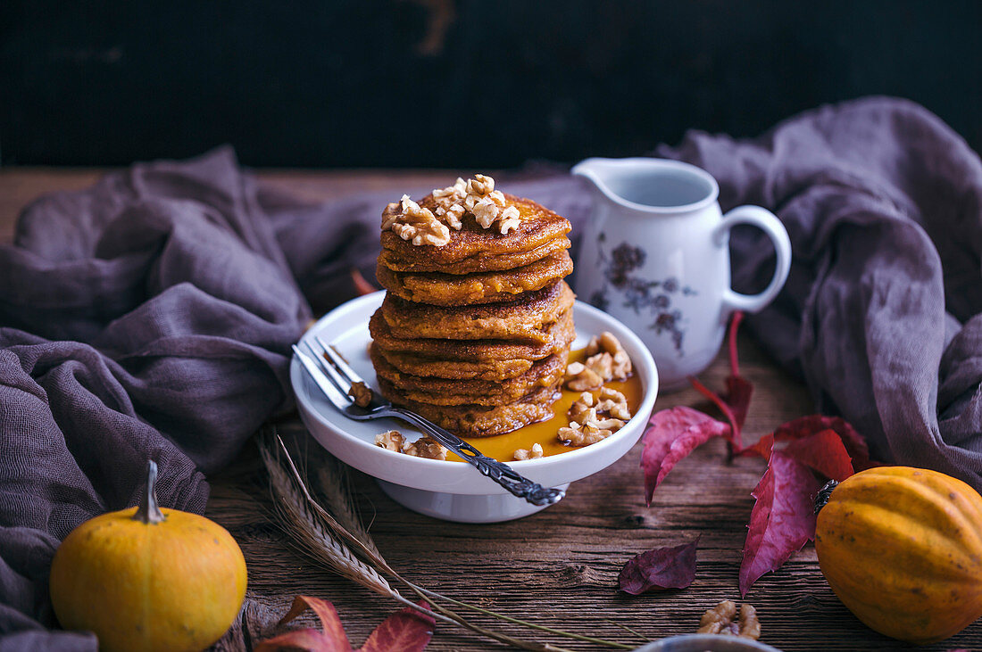 Pumpkin pancakes topped with walnuts and maple syrup served on a little cake stand on rustic wooden table