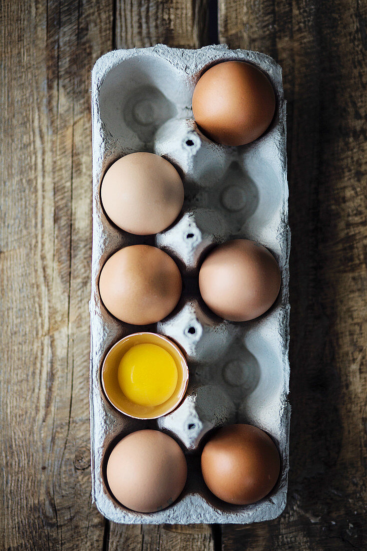 Ganze braune Eier und ein aufgeschlagenes Ei in Eierkarton (Aufsicht)