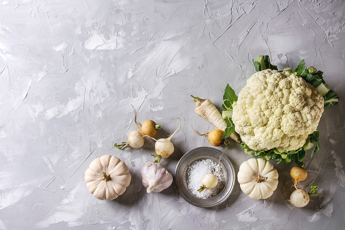 Variety of white vegetables raw organic cauliflower, pumpkins, garlic, parsnip and radish with plate of sea salt over gray texture background