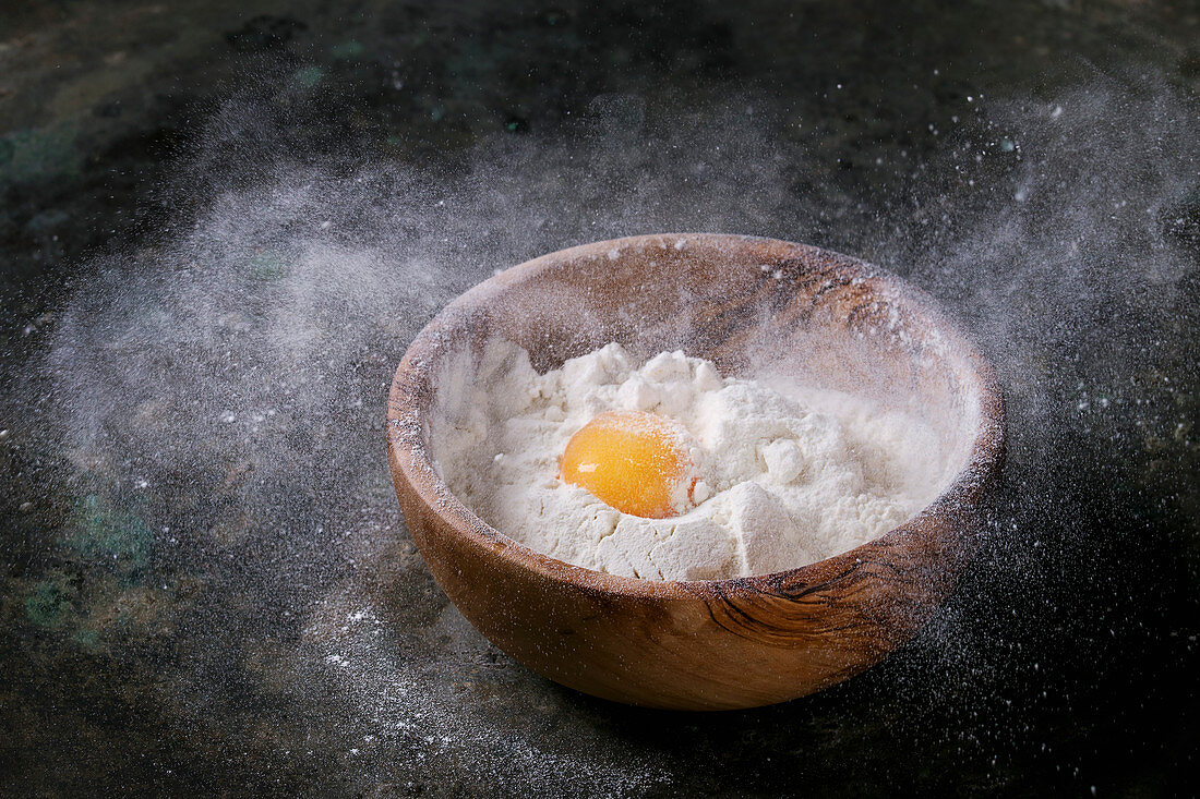 Olive wood bowl with wheat flour and whole egg yolk over old dark metal background