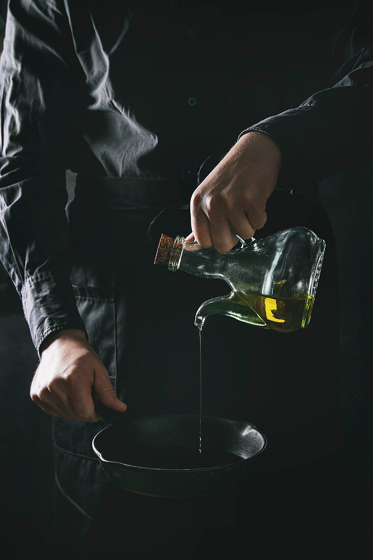 Mann giesst Olivenöl aus Glaskaraffe in Gusseisenpfanne