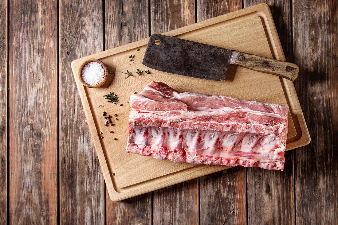 Fresh raw uncooked whole rack of pork loin with ribs on wooden cutting board with salt, thyme and butcher clever over old wood plank table