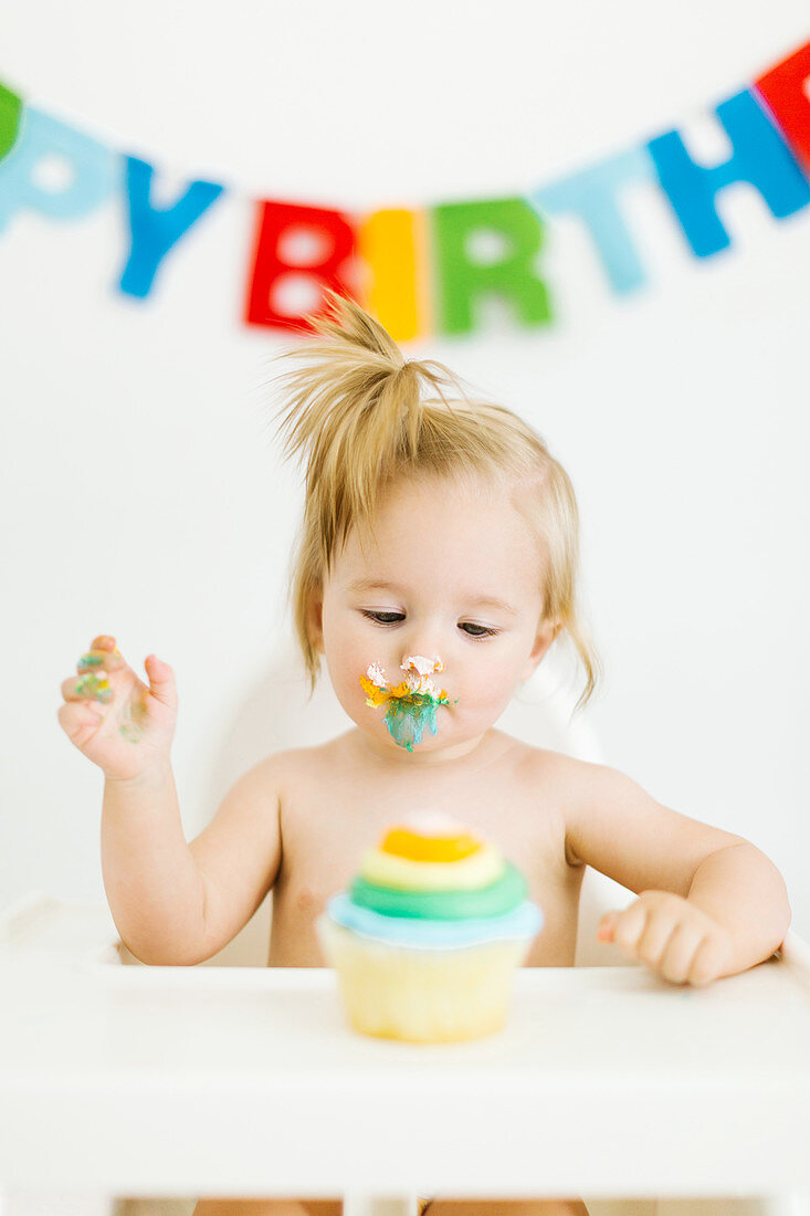 Kleines Mädchen sitzt im Hochstuhl und isst Muffin zum ersten Geburtstag