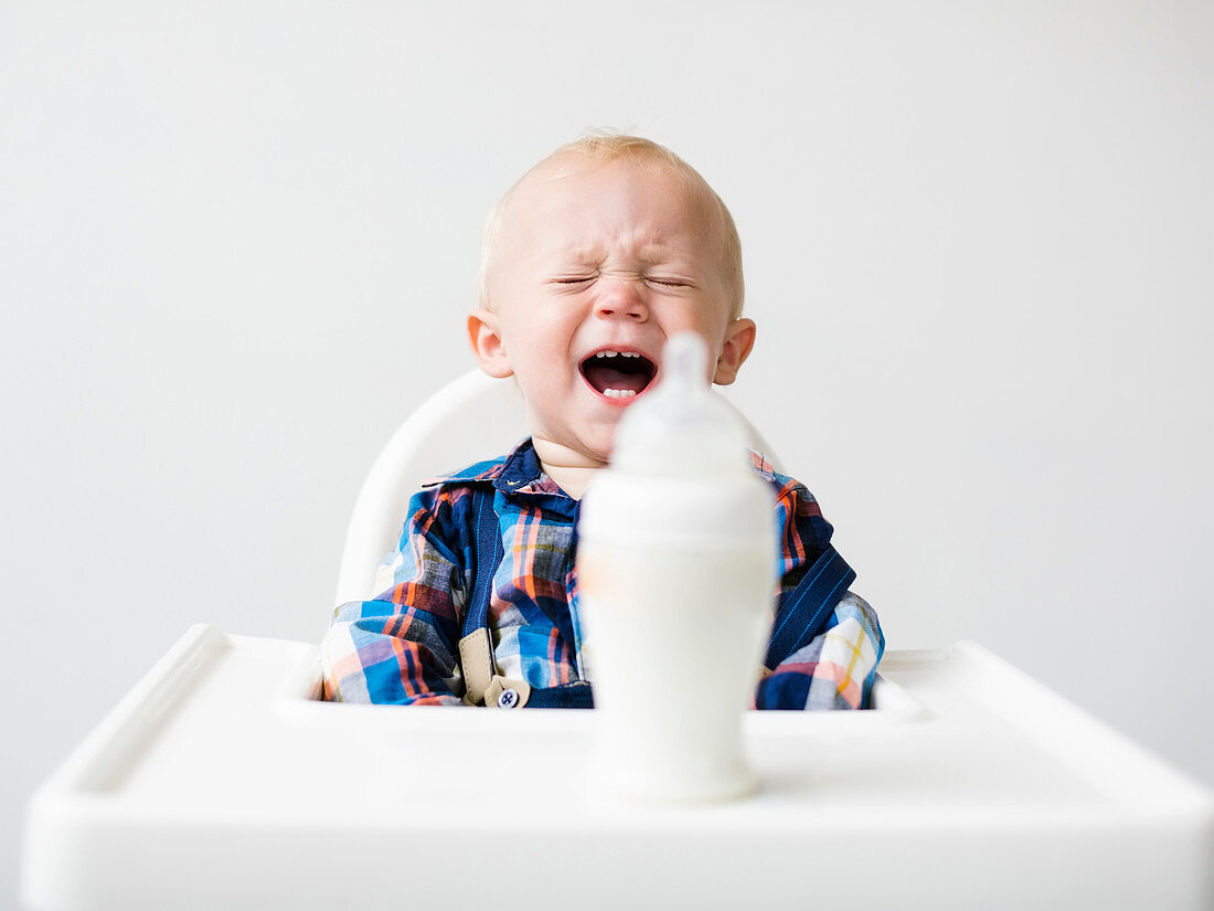 A little boy crying in a highchair in front of a milk bottle