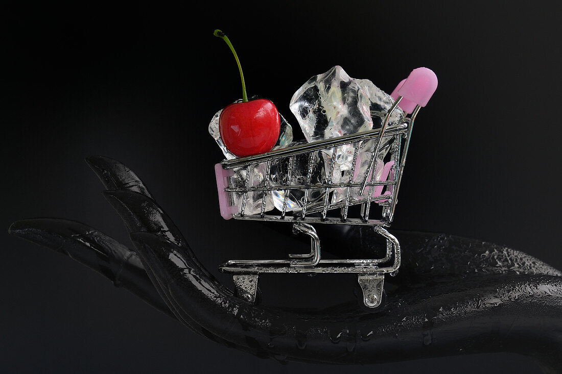 A black hand holding a mini shopping cart filled with ice cubes and a cherry