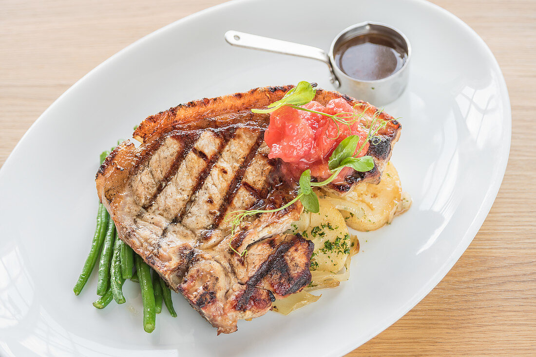 Grilled steak with long green beans, potatoes and grilled tomatoes with steak sauce on a white plate and light wooden background