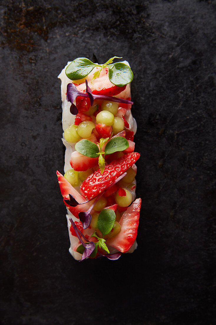 Seebarschfilet mit Erdbeeren und Melonenbällchen vor schwarzem Hintergrund