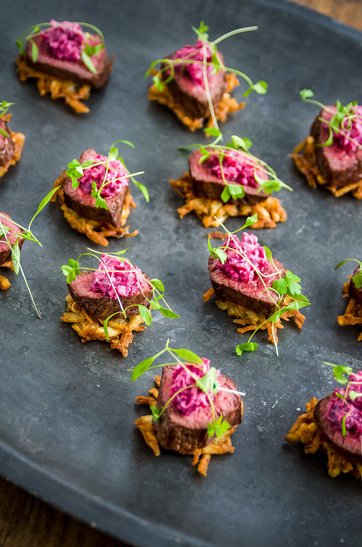 Canapes: crispy potato hash, venison and beetroot salad garnished with parsley leaves on a dark metal serving plate