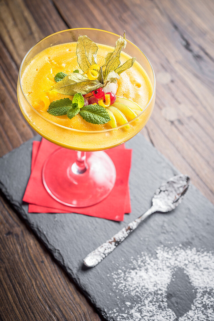 Mango sorbet decorated with citrus peel, mint, redcurrant in a glass with a red napkin and a spoon on a slate board and wooden background