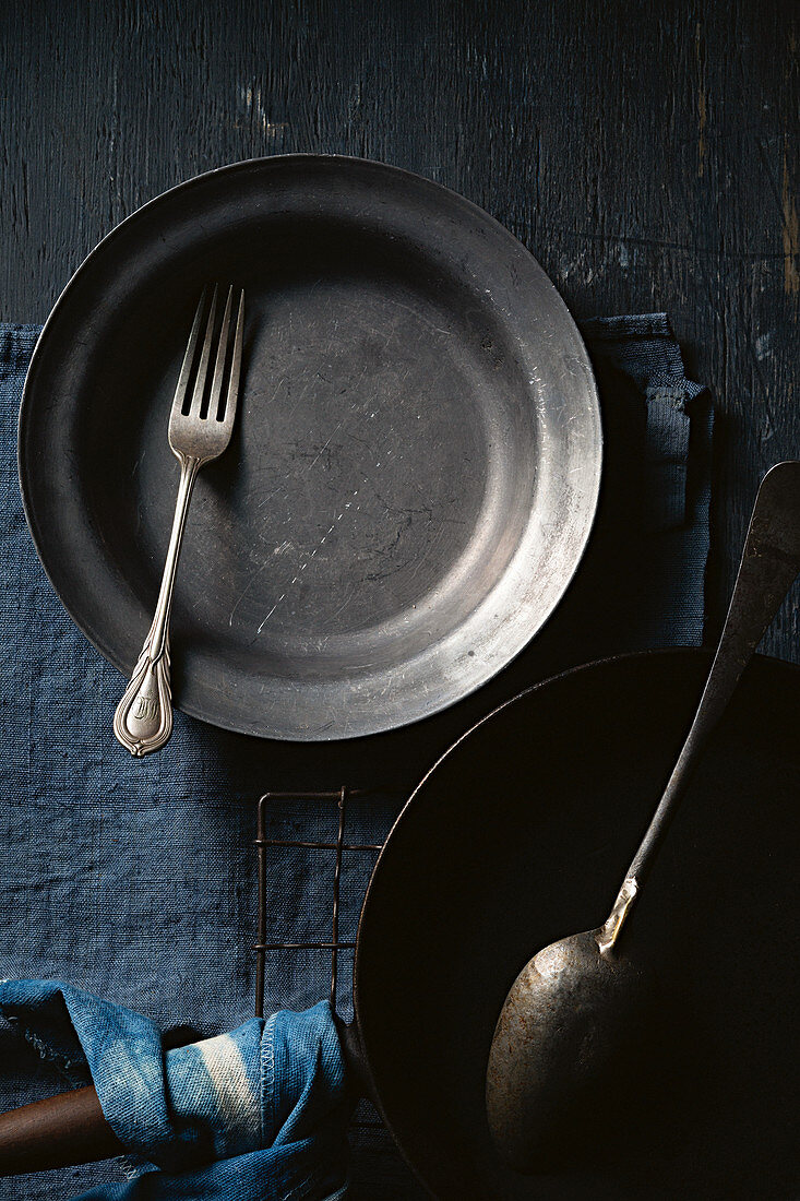 Schwarzer Teller mit Vintage Gabel, Pfanne und Löffel auf blauem Untergrund