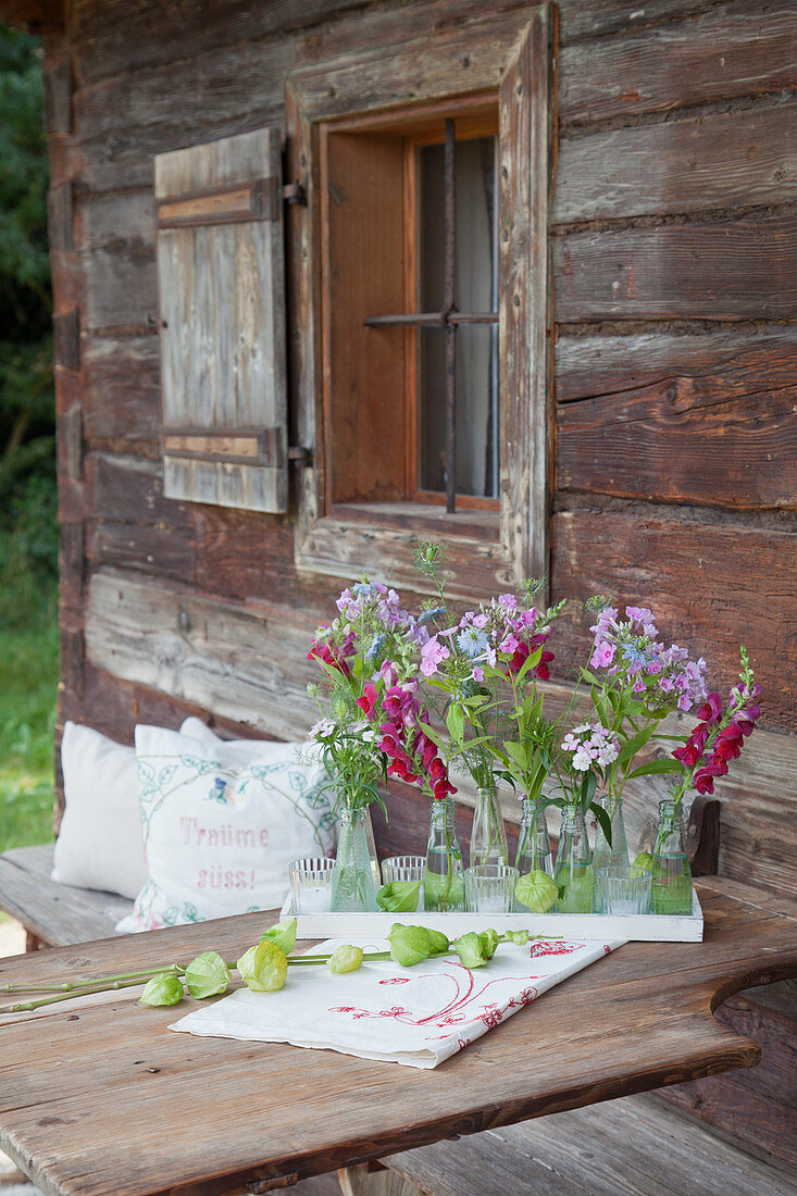 Kleine Flaschen mit Phlox, Jungfer im Grünen und Löwenmäulchen als Tischdekoration vor rustikalem Bauernhaus