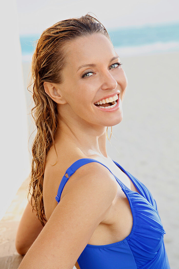 A blonde woman with wet hair on a beach wearing a blue bathing suit