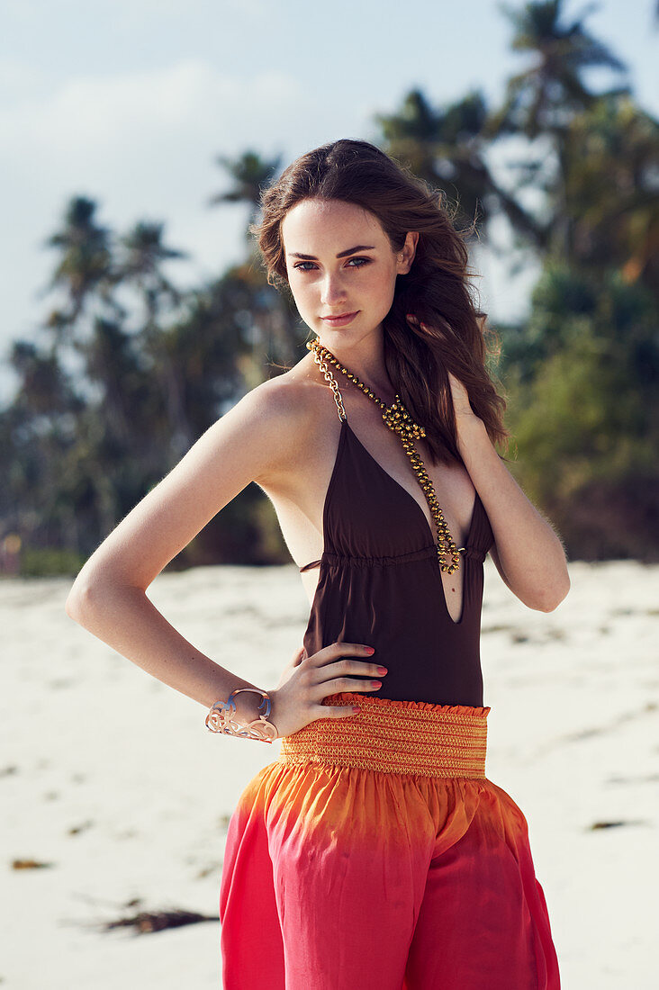 A young brunette woman on a beach wearing a brown bathing suit and harem trousers