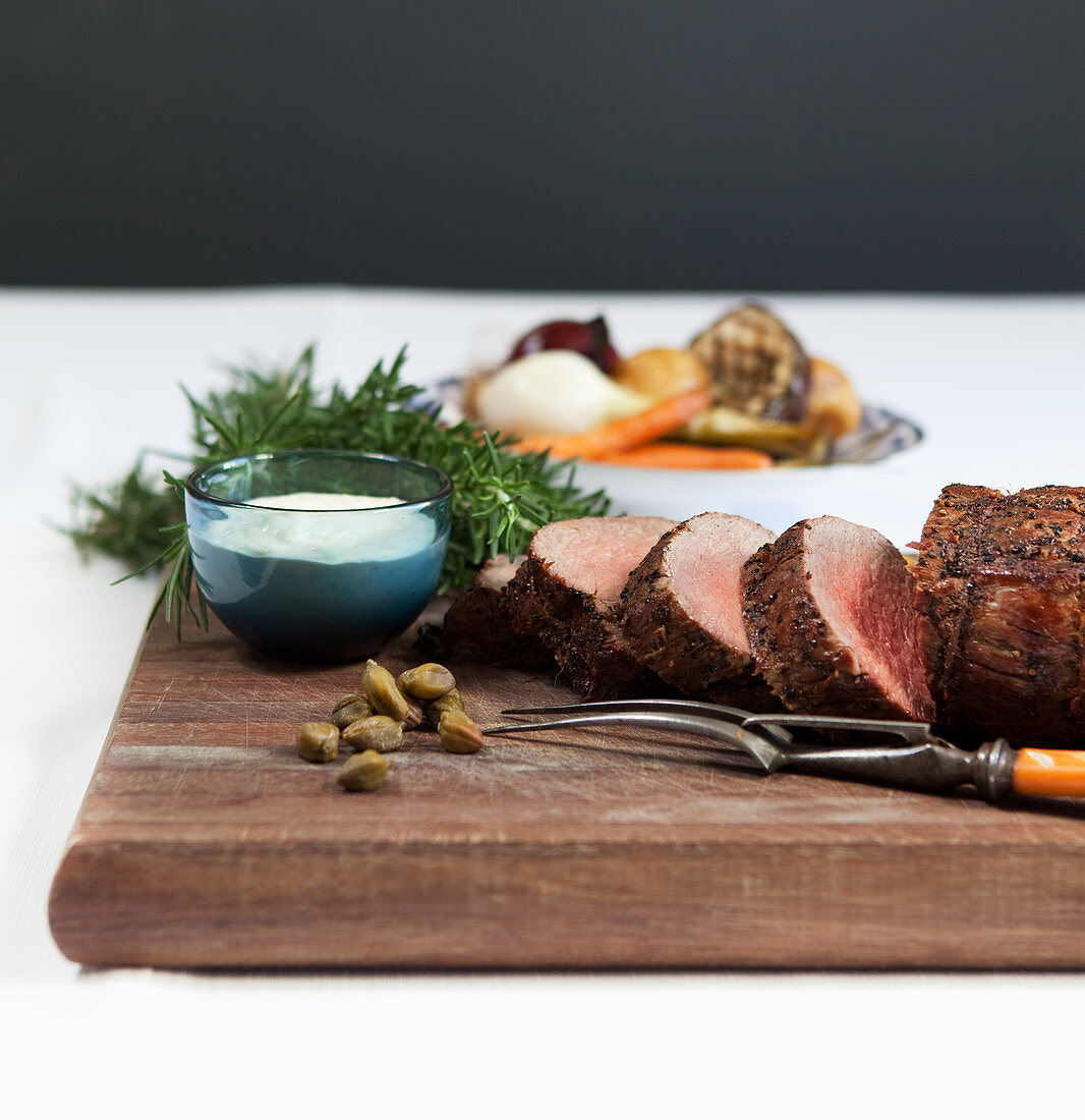 Rolled roast beef with horseradish sauce, rosemary and capers, roast vegetables in the background