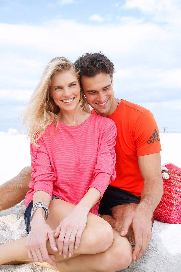 Blonde Frau in rosa Bluse und brünetter Mann in orangefarbenem T-Shirt sitzen am Strand