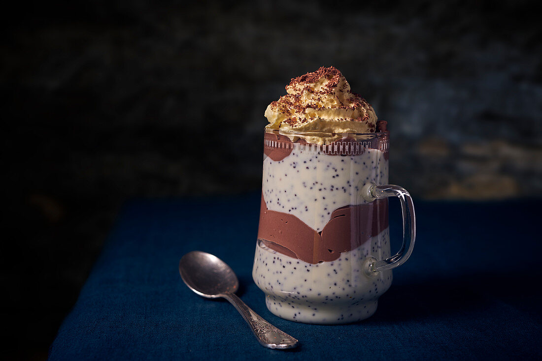 A layered dessert with poppy seeds and chocolate cream in a tall glass with a handle