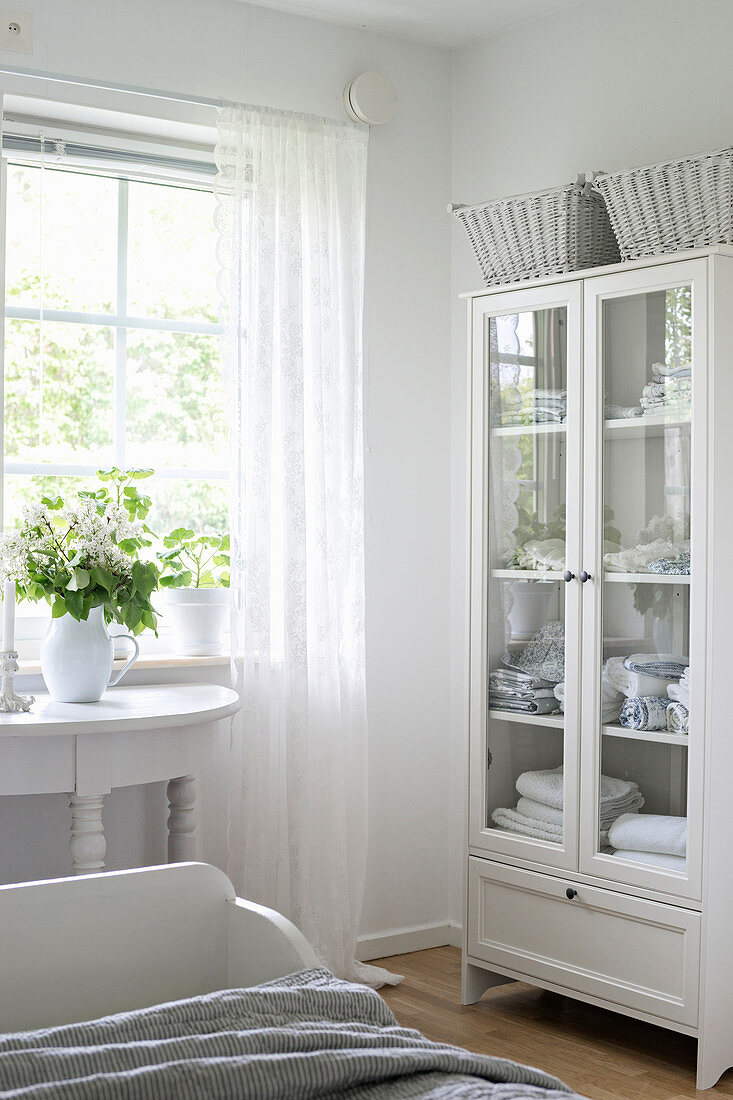 Linen in glass-fronted cabinet in bedroom decorated entirely in white