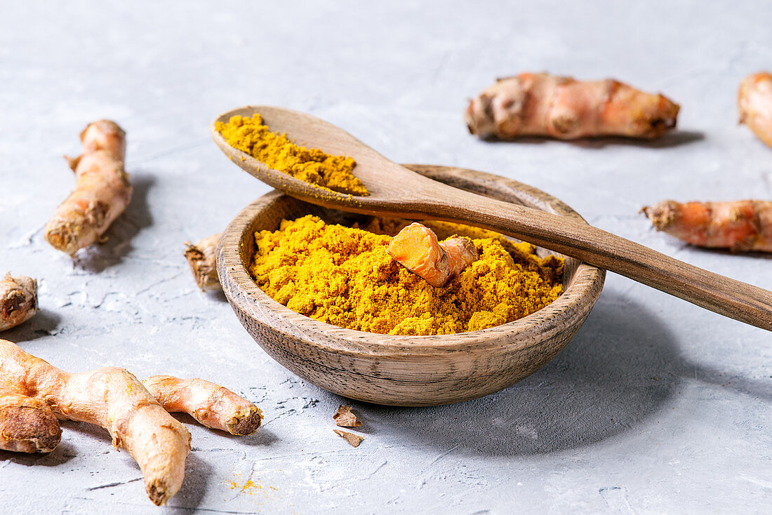 Turmeric roots and powder in wooden bowl over gray concrete background