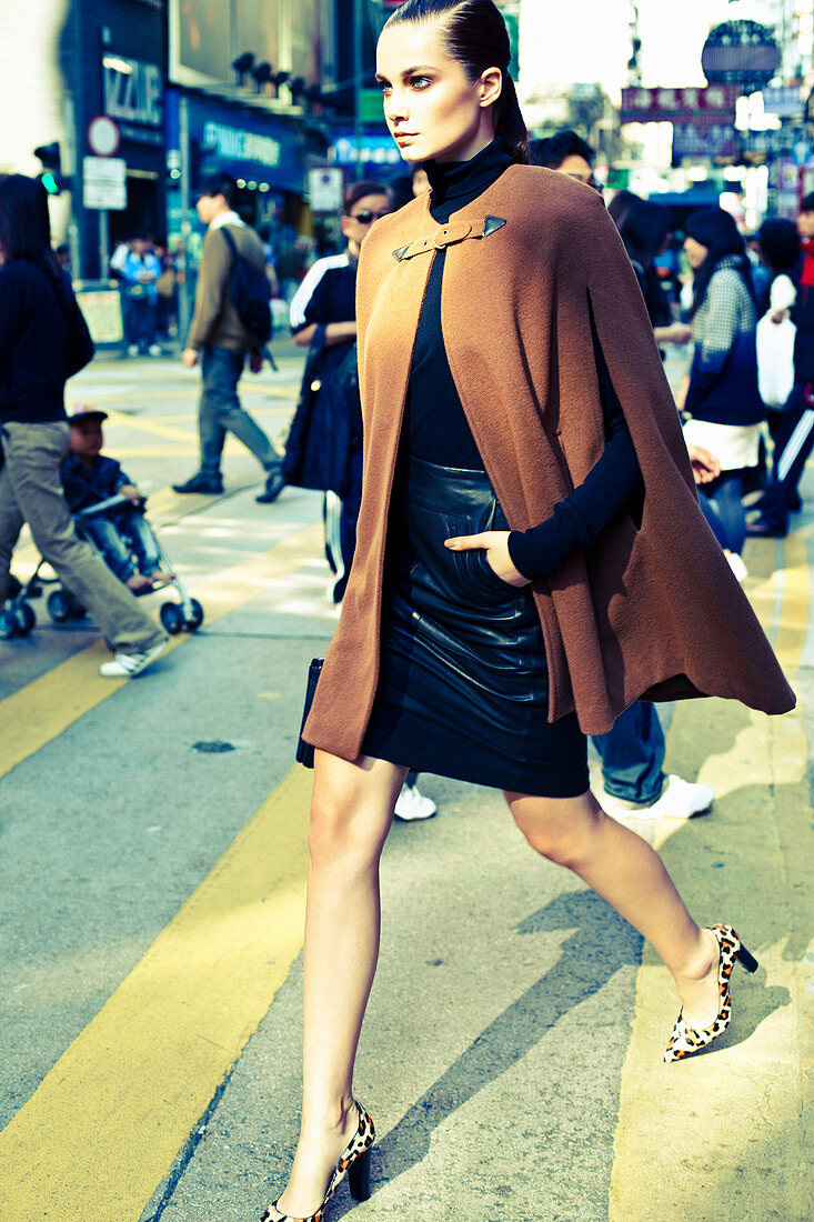 A young woman walking across a road wearing a brown cape, a black turtleneck jumper and a black skirt