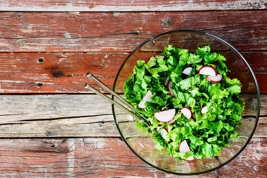 Lettuce with radishes in a glass bowl on a wooden background (top view)