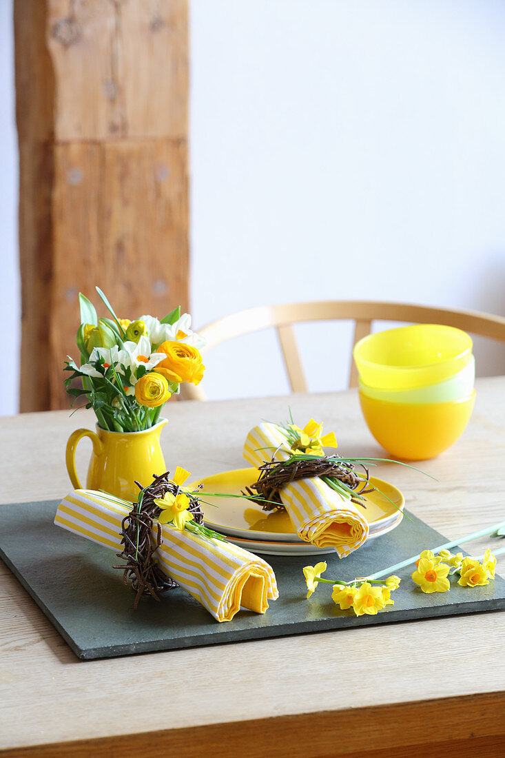 Gelber Teller, und gelb-weiß gestreifte Serviette mit Osterkränzchen und kleiner Blumenstrauß in Krugvase