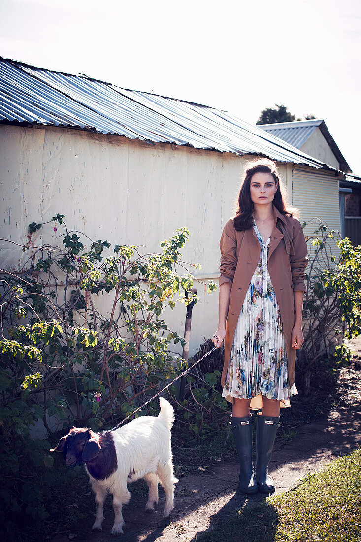 Dunkelhaarige Frau im Sommerkleid, Trenchcoat und Gummistiefeln mit Hund