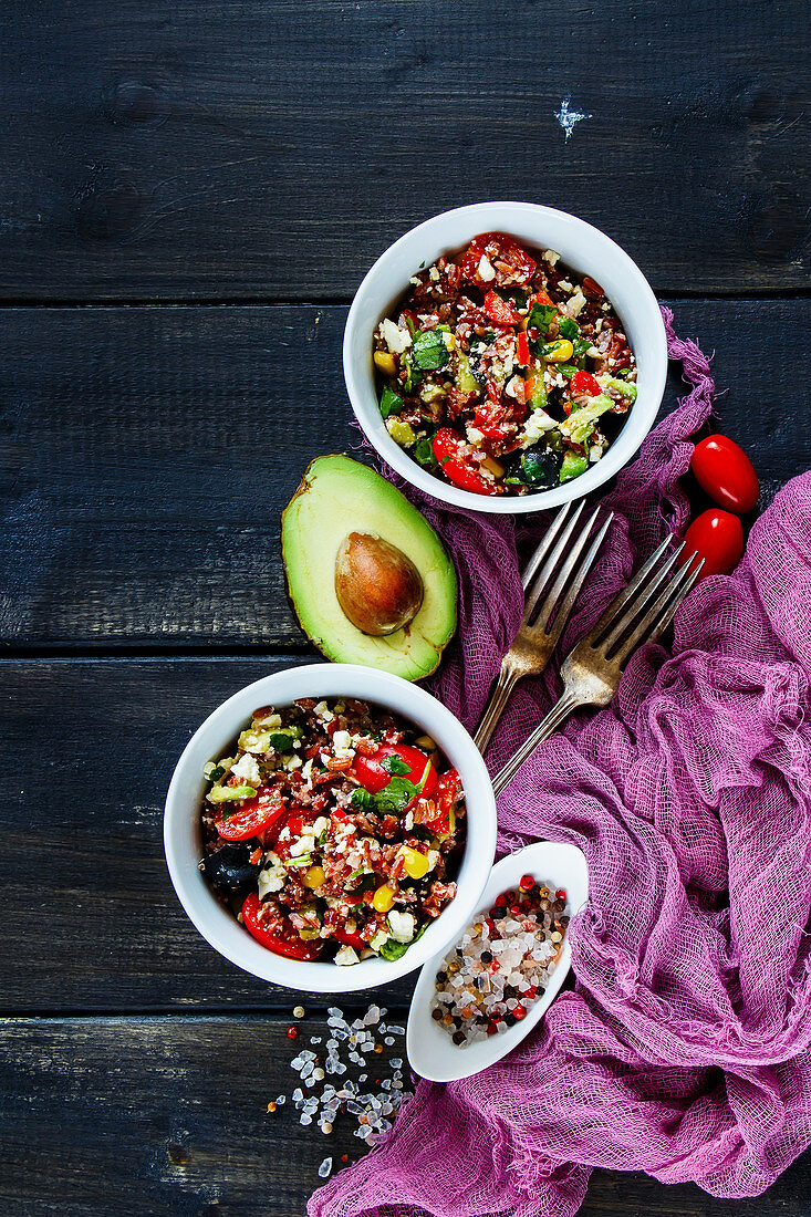 Homemade red rice salad with feta cheese, cherry tomatoes, avocado, black olives and ingredients on wooden background