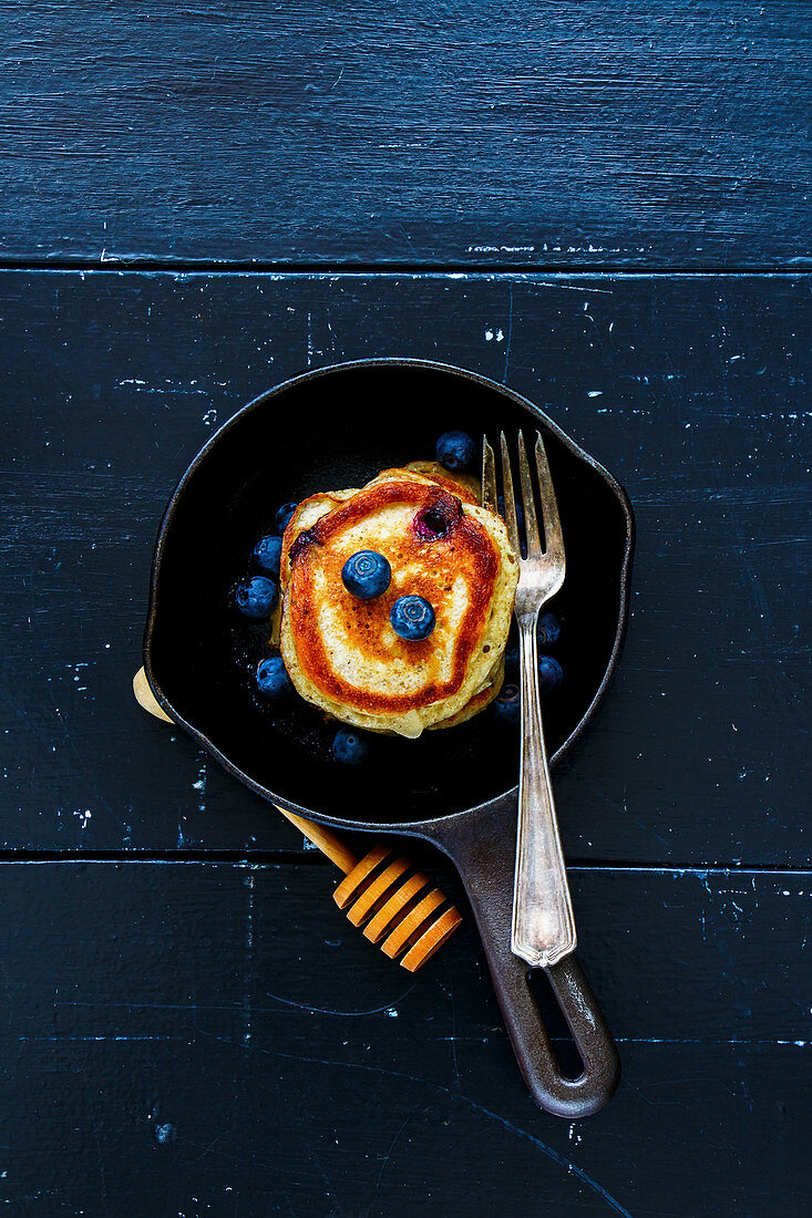Top view of blueberries homemade pancakes with honey and fresh berries for breakfast in vintage cast iron pan over rustic wooden background