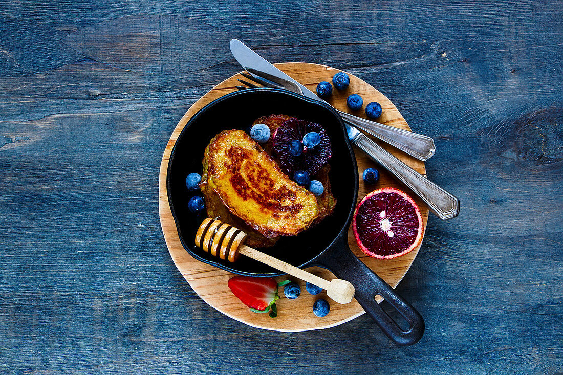 Homemade Cinnamon French Toasts with honey, berries and bloody orange in vintage cast iron pan for breakfast on rustic wooden background