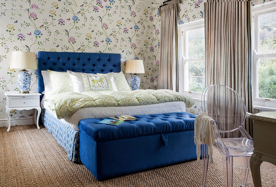 Double bed with royal-blue headboard and ottoman in bedroom with floral wallpaper