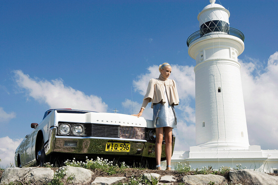 A blonde woman wearing a silver dress and a naturally white jacket standing by a car