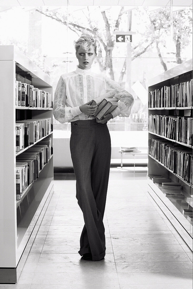 A blonde woman wearing dark trousers and an elegant white embroidered blouse in a library (black-and-white shot)