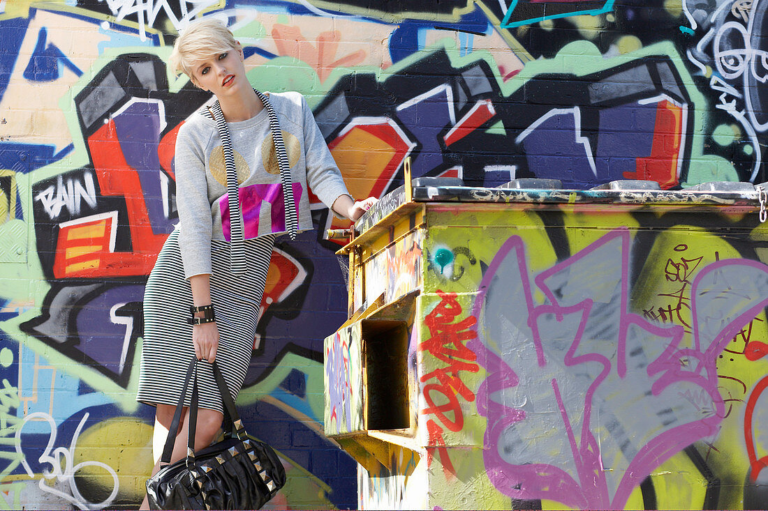 A blonde woman wearing a grey jumper and a skirt in front of a graffitied wall