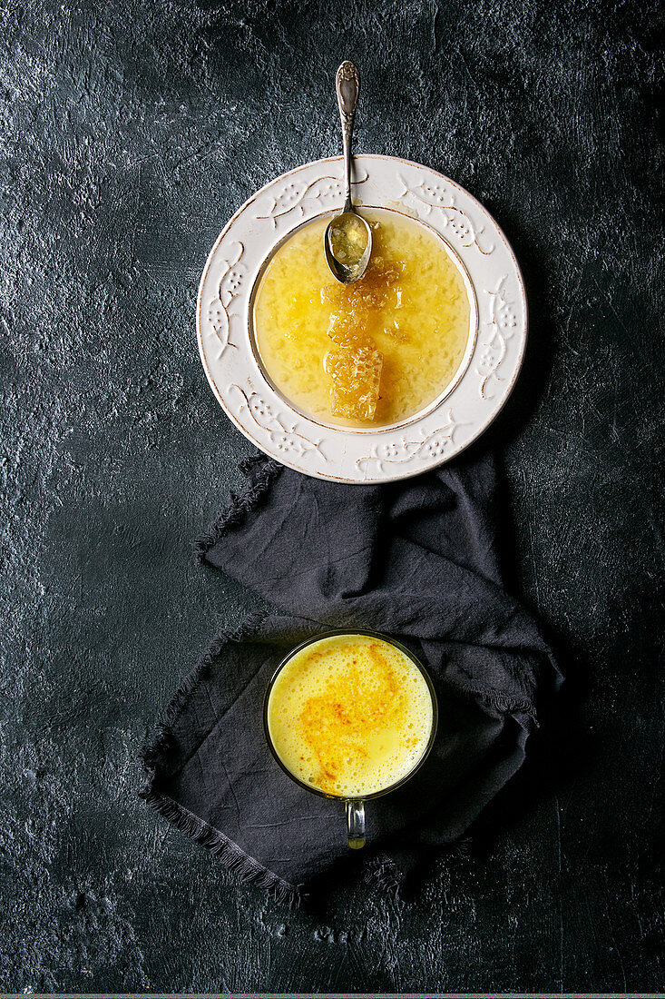 Honeycombs in liquid honey on white vintage plate and cup of turmeric latte on cloth over black texture background