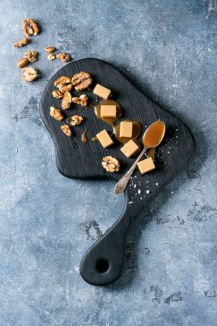 Salted caramel fudge candy served on black wooden board with fleur de sel, caramel sauce and caramelized walnuts