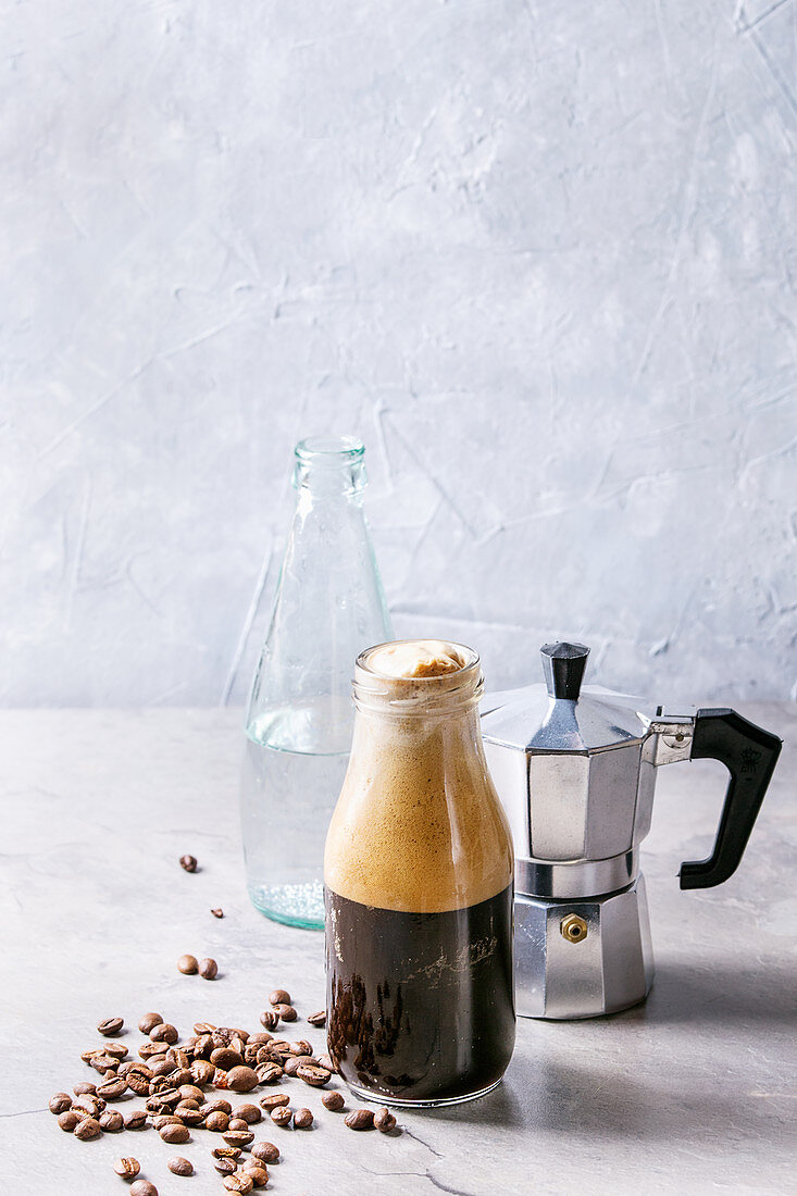 Bottle of fizzy iced Coffee espresso with bottle of sparkling water, coffee maker and roasted beans over grey table