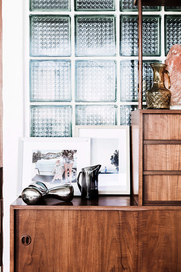 Retro sideboard with decorative objects and framed photos in front of a glass brick wall