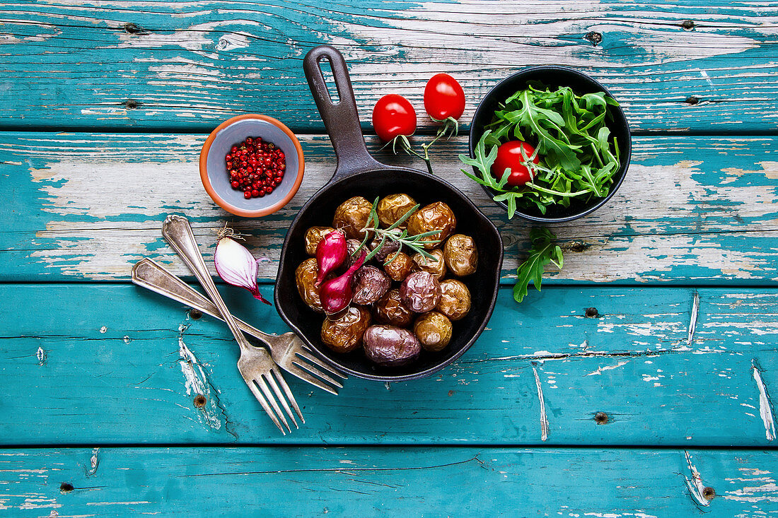 Fried baby potatoes in old cast iron pan and salad with tomato and arugula in bowl on turquoise wooden background