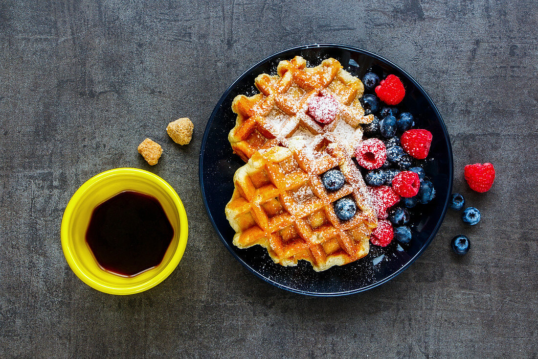 Homemade Belgian soft waffles with berry on black plate, espresso cup and cubes of brown sugar