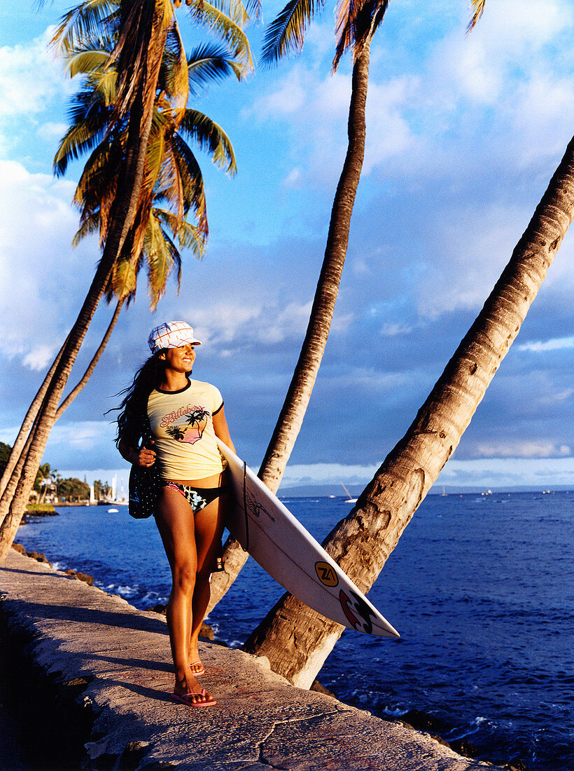A brunette woman wearing a cap, a t-shirt and bikini trousers with a surfboard by the sea
