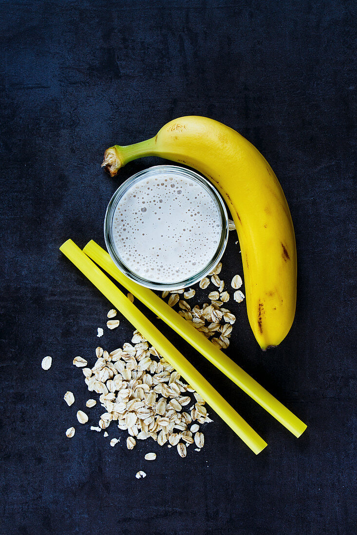 A banana smoothie in a glass next to oats, straws and a banana